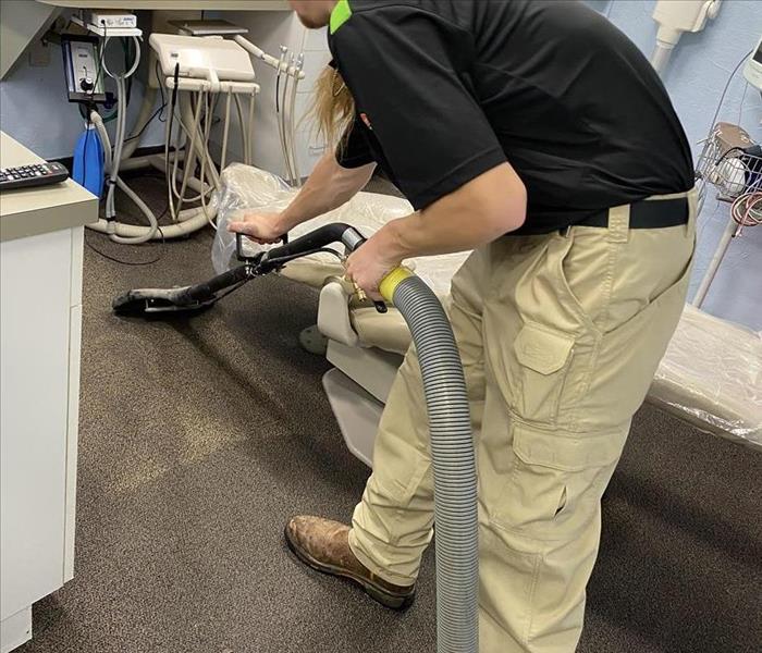 Technician in black polo and khaki pants using a vaccum-like drying tool to remove water for carpet in a dentist office.