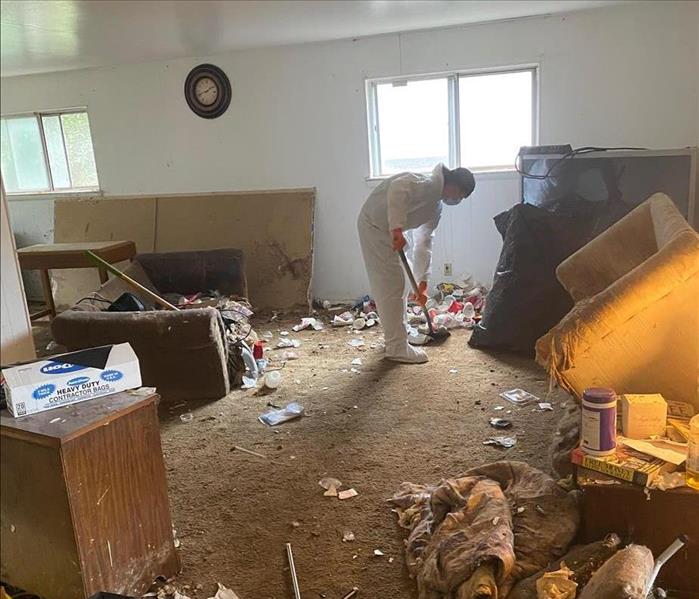 Man in personal protective wear shoveling trash out of a carpeted living room.