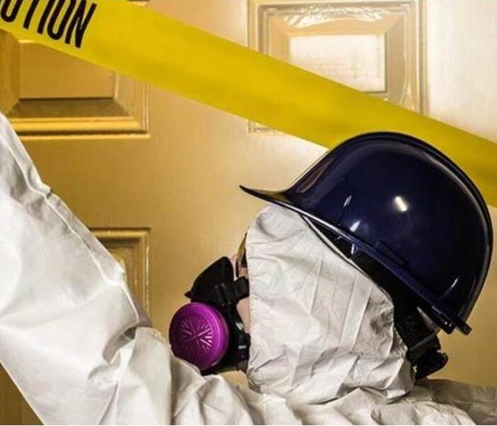 Worker in personal protective equipment placing yellow "caution" tape across a doorway.